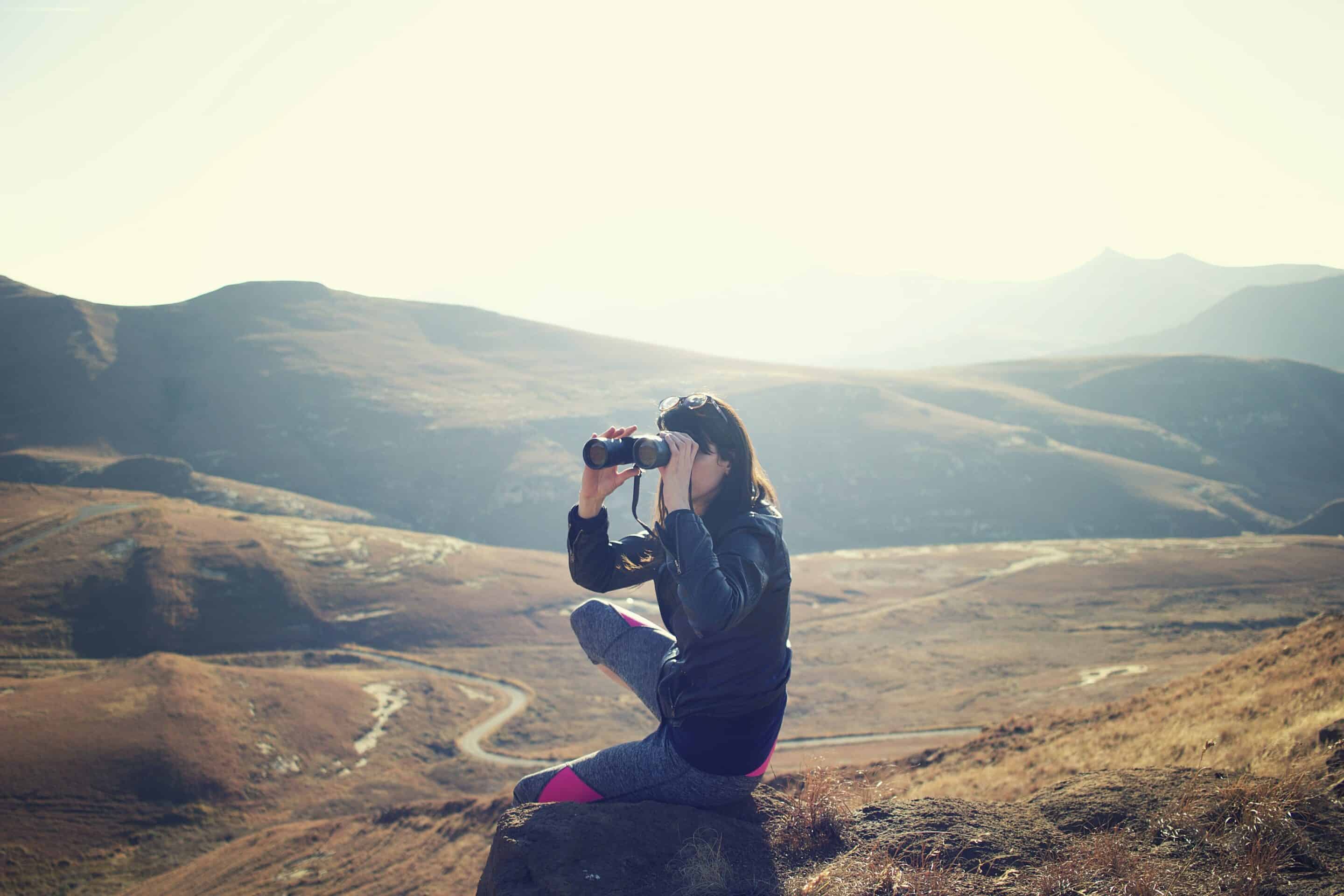 a woman crouching on a mountain looking into binoculars