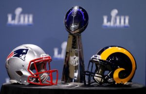 Patriots helmet and rams helmet sit on a table facing each other in front of superbowl trophy. 
